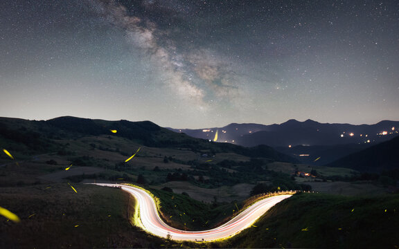 View of the Milky Way galaxy over a winding mountain road with car light trails and fireflies in the dark landscape of Albareto, Emilia-Romagna, Italy.