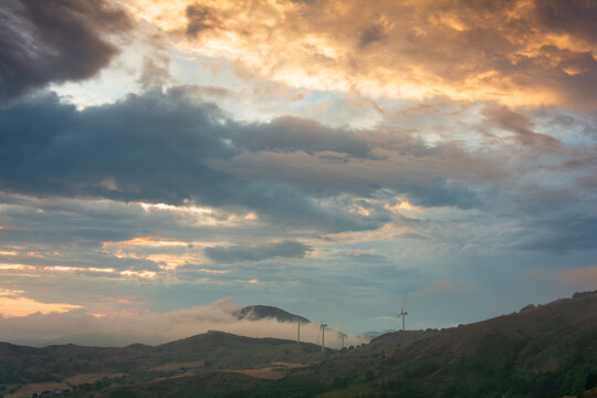View of wind turbines of Parco Eolico Passo Cappelletta on rolling hills under a dramatic sunset sky with mist Albareto, Emilia-Romagna, Italy.
