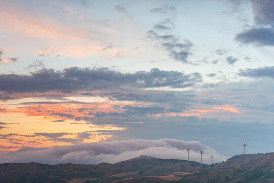 View of Parco Eolico Passo Cappelletta wind turbines on rolling hills under a sunset sky with soft clouds and orange light Albareto, Emilia-Romagna, Italy.