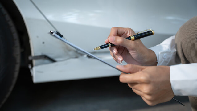 Close-up of person holding pen and clipboard inspecting white car damage for insurance claim or repair estimate in daylight.