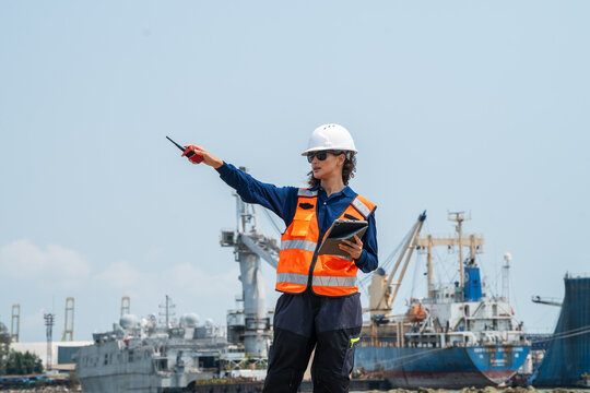 Woman wears a white hard hat and orange safety vest. She points with a pen while holding a tablet. woman engineer communicating via radio while inspecting maritime logistics at an industrial harbor.