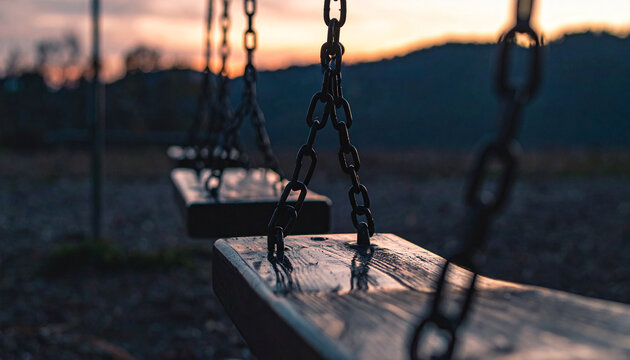 Empty wooden swings with metal chains hang in a park at dusk with a soft orange sunset sky
