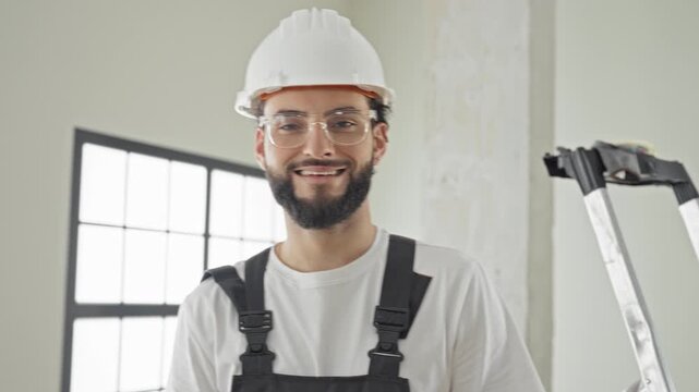 Man construction worker wearing hardhat, safety glasses and apron smiles while holding a ladder inside a renovated building; pride professionalism.