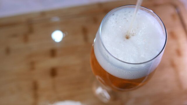 POV of someone filling a glass of beer on a wooden table. Close-up of a person pouring beer into a glass. Close-up of bubbles bursting in a thick beer foam. Detail of a thick head of beer. A beverage
