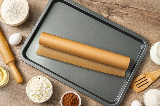 Dark gray baking sheet holds rolled parchment paper. Surrounding items flour, eggs, cacoa powder, rolling pin, wooden spoon. wooden surface adds rustic charm. view from above