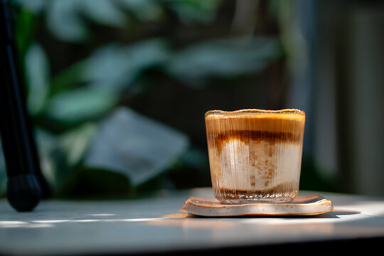 A layered dirty coffee in a clear glass sits on a ceramic coaster, lit by soft sunlight. Blurred tea and water in the background with lush green leaves create a calm, natural cafe mood.