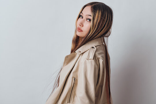 Young woman posing in beige leather jacket on light background looking sideways with long straight hair and casual style for fashion and portrait photography.