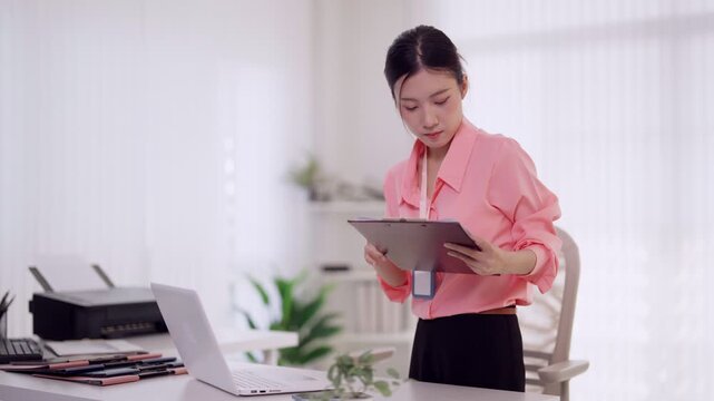 Asian businesswoman reviewing documents standing at office desk using laptop computer multitask professional entrepreneur working at modern workplace checking