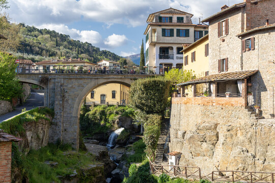 View of the historic stone bridge and waterfall surrounded by traditional buildings and lush green hills in Loro Ciuffenna, Tuscany, Italy.