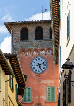 View of the historic clock tower with its large white face and colorful coats of arms framed by narrow village streets in Loro Ciuffenna, Tuscany, Italy.