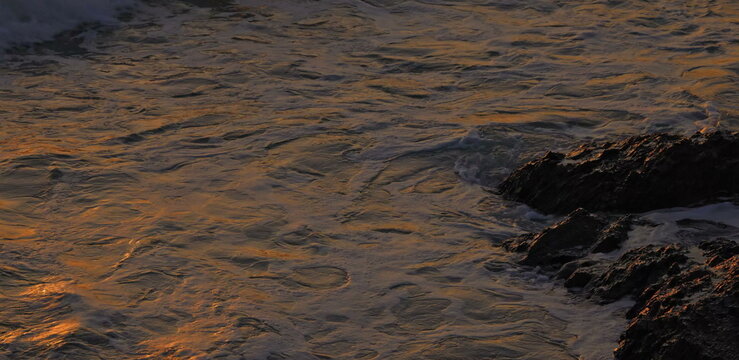 Golden sunset light reflecting on ocean waves and dark jagged rocks