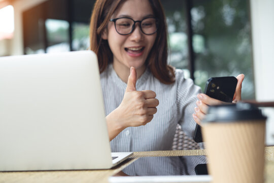 Asian woman happy video call using mobile phone and showing thumb up during online working at home. Successful business woman celebrating on video chat using smartphone at outdoor cafe