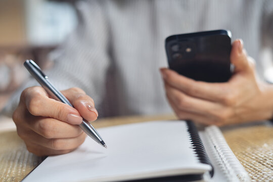 Woman hand with a pen writing on notebook and using mobile phone searching the information, online research, business planning 