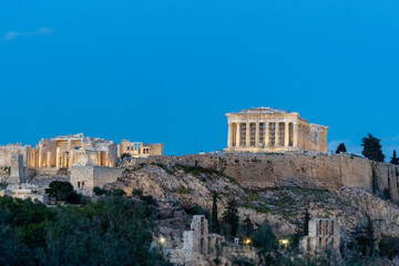 Naklejka premium A majestic evening view of the illuminated Acropolis of Athens, featuring the Parthenon and the Odeon of Herodes Atticus under a deep blue twilight sky, as seen from Philopappos Hill.