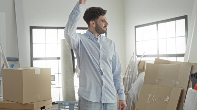 Young hispanic man with moving boxes rubbing his neck and smiling inside a building with stacked cartons and ladder visible; relief settling transition.