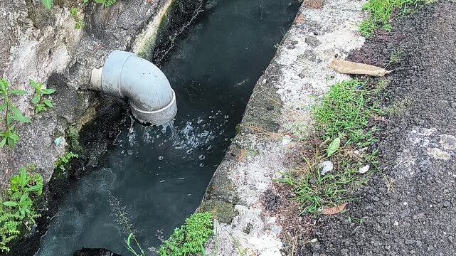 A grey pipe discharges dark, murky water into a concrete drainage ditch with green vegetation on its banks, indicating water pollution.
