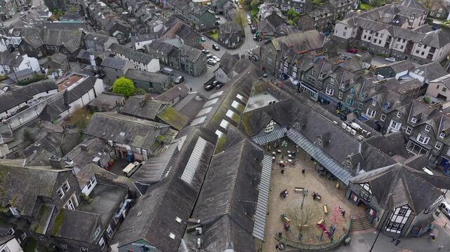 A rising drone shot reveals the slate rooftops and market square of Ambleside town centre, Cumbria, with the covered market hall and surrounding shops clearly visible.