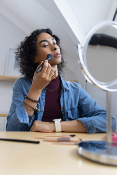 Non-binary person applying makeup with brush at vanity table using round mirror wearing denim top
