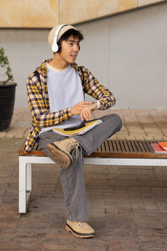 Asian teen student sitting on campus bench wearing beanie and headset, holding notebooks and phone