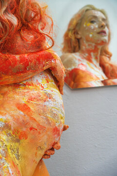 Pregnant woman covered in vivid paint holds belly while looking at her reflection creating expressive artistic scene with strong color texture and emotional maternal focus