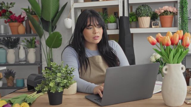 Woman florist in apron raises fists over laptop in studio, smiling amid tulips and plants; small business joy.