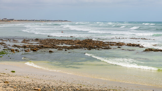 Dramatic Brittany coastline near Audierne and Plouhinec, with crashing Atlantic waves, rugged cliffs, turquoise water, coastal vegetation, and distant seaside houses on a windy surf day.