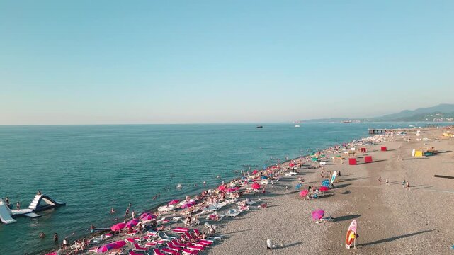 Aerial fly over view busy pebble beach in Batumi, Georgia. Rows of pink umbrellas, sunbathers, and calm Black Sea waters on clear summer evening at sunset.