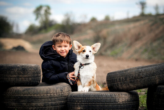 Smiling boy and Corgi dog sitting among old car tires outdoors.