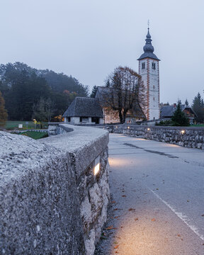 View of the historic Church of St. John the Baptist and a stone bridge with glowing lights at dusk in Municipality of Bohinj, Radovljica, Slovenia.