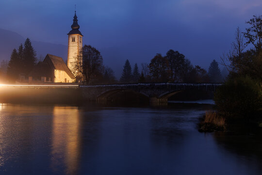 View of the illuminated Church of St. John the Baptist and the stone bridge reflecting in the calm waters of Lake Bohinj at twilight, Municipality of Bohinj, Radovljica, Slovenia.