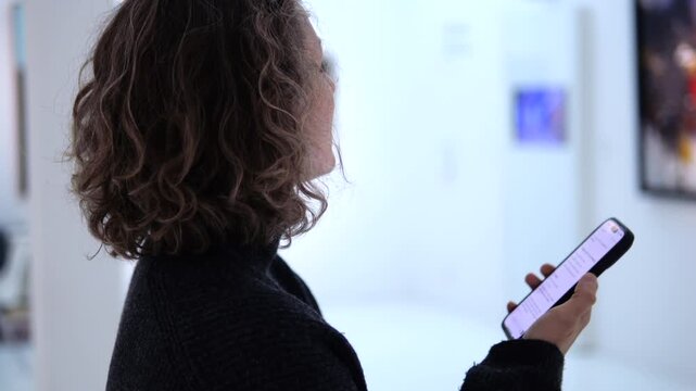 Adult woman with curly hair using her smartphone for an audio guide while visiting a modern art gallery, looking at pictures and exploring the exhibition with technology
