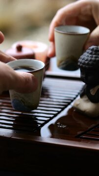 Close up view of hands participating in a traditional chinese tea ceremony, picking up steaming ceramic cups from a wet wooden tray with a small buddha statue and clay teapot nearby