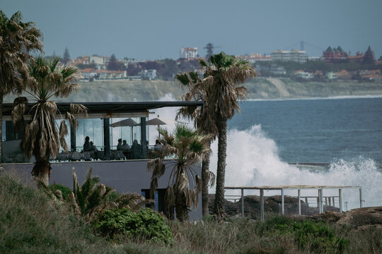 View of a seaside restaurant terrace with people dining behind glass walls as large waves crash against the rocky shore near palm trees in Cascais, Lisbon, Portugal.