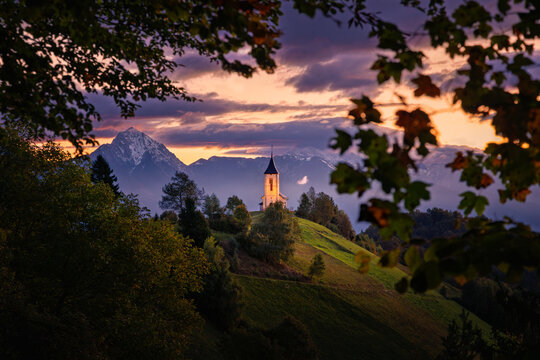 View of the Church of St. Primus and Felician on a grassy hill with the Julian Alps under a sunset sky Jamnik, Kranj, Slovenia.