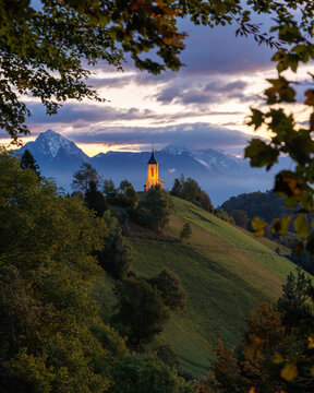 View of Jamnik Church on a grassy hilltop with snow-capped mountains under a dramatic sunset sky framed by leaves Jamnik, Kranj, Slovenia.