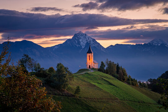 View of the Church of St. Primus and Felician on a grassy hill against the backdrop of snow-capped mountains during sunset in Jamnik, Kranj, Slovenia.