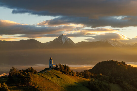 View of Jamnik Church on a grassy hilltop with the Julian Alps and morning mist under a golden sunrise sky Jamnik, Kranj, Slovenia.