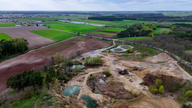 Aerial photograph of an active sand and gravel pit with machinery, surrounded by cultivated fields, green forests, and a rural village, under a partly cloudy sky, showing several small post-mining pon