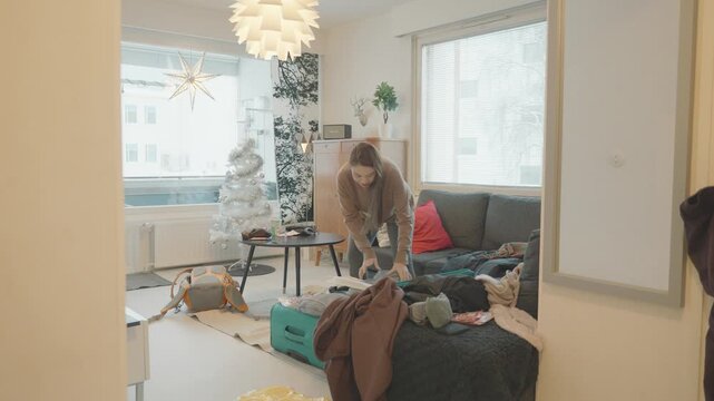 A woman tidies up her living room filled with clothes and decorations, creating a warm and inviting atmosphere. The festive tree and cozy furniture contribute to a comfortable home environment.