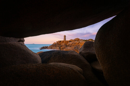 View of Ploumanac'h Lighthouse framed by massive pink granite boulders along the rocky coastline under a soft twilight sky in Perros-Guirec, Bretagne, France.