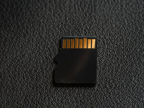 A detailed close-up shot focusing on the connector side of a microSD card. The high-contrast lighting highlights the metallic texture of the gold pins .