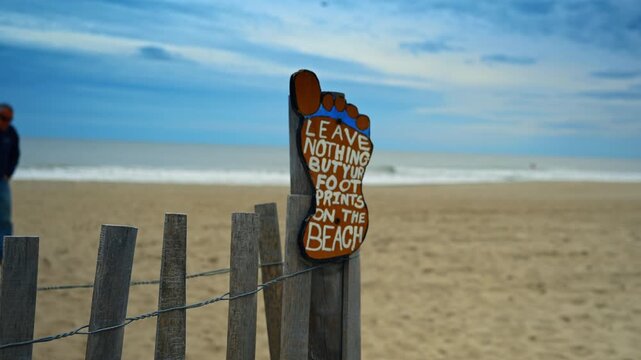 Wooden foot shaped sign with environmental message on beach. A sign on a wooden fence saying Leave nothing but your footprints on the beach against an ocean background.