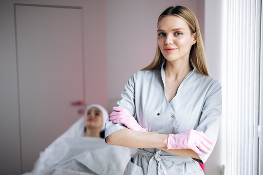 Medical aesthetician with crossed arms wearing gloves in a beauty clinic, client relaxing on bed in background