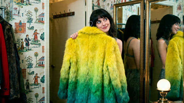 [Young woman] [with dreamy mood] [trying on a bright gradient fur coat and dreaming of the purchase in a fitting room]