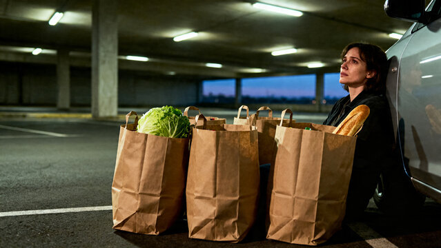 [Young woman] [with pensive mood] [sitting on the parking lot floor, leaning against a car, with grocery bags in front of her]
