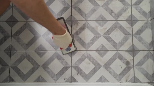 Close up of construction worker applying grey grout to patterned ceramic tiles on a bathroom wall. Tiler uses grout float to finish wall above bathtub during renovation