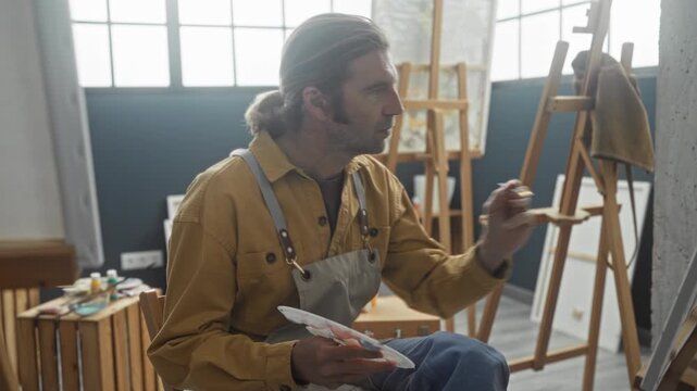 Man painting with brush on canvas in studio, wearing apron and suspenders while reaching with his hand to make brushstrokes on easel; concentration.