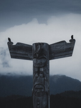 View of a traditional wooden totem pole with intricate carvings and two black birds perched on its wings against misty mountains in Canada.