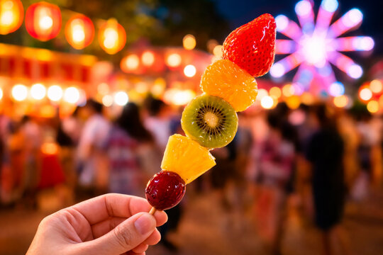 Candied fruit skewer (tanghulu) featuring a strawberry, mandarin orange, kiwi, pineapple, and grape at a vibrant night festival.