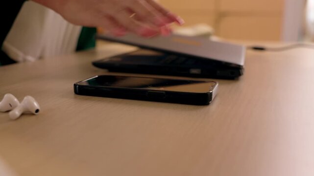 Laptop hand desk smartphone earbud office workspace wooden technology indoor scene, hands typing laptop with smartphone sliding beside it, wireless earbud nearby, warm light modern remote work setup